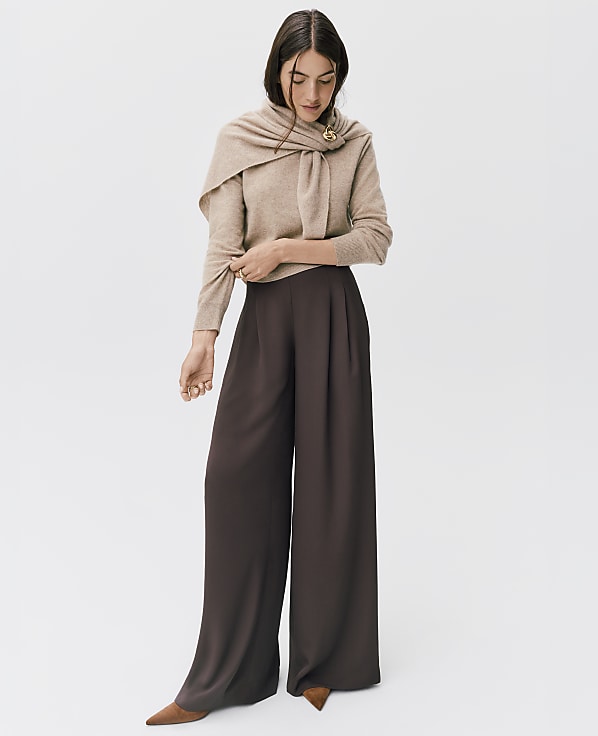 A woman wearing a beige sweater with a matching scarf and wide-leg brown trousers stands against a plain white background.