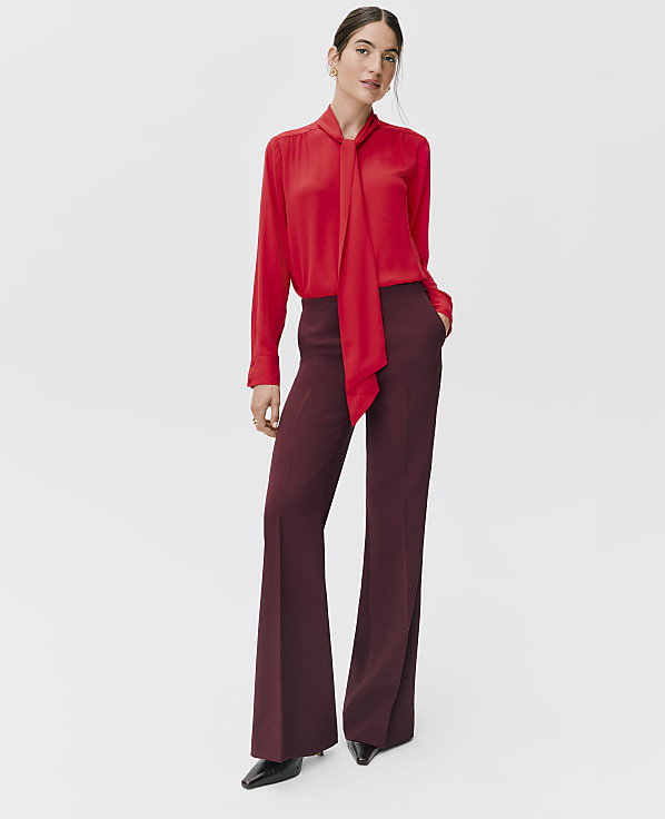 A woman wearing a red pussy-bow blouse and wide-leg burgundy trousers stands against a plain white background.
