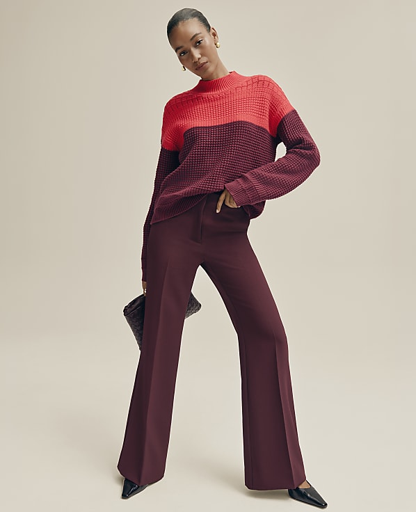 A woman wearing a red and burgundy color-blocked sweater and matching wide-leg trousers stands against a plain, neutral-toned background.
