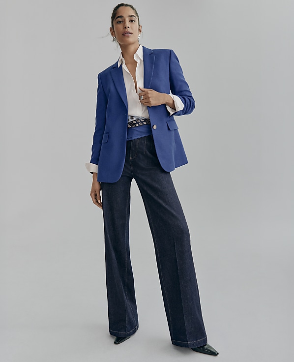 A woman wearing a blue blazer, white shirt, and wide-leg dark denim jeans stands against a plain light gray background.