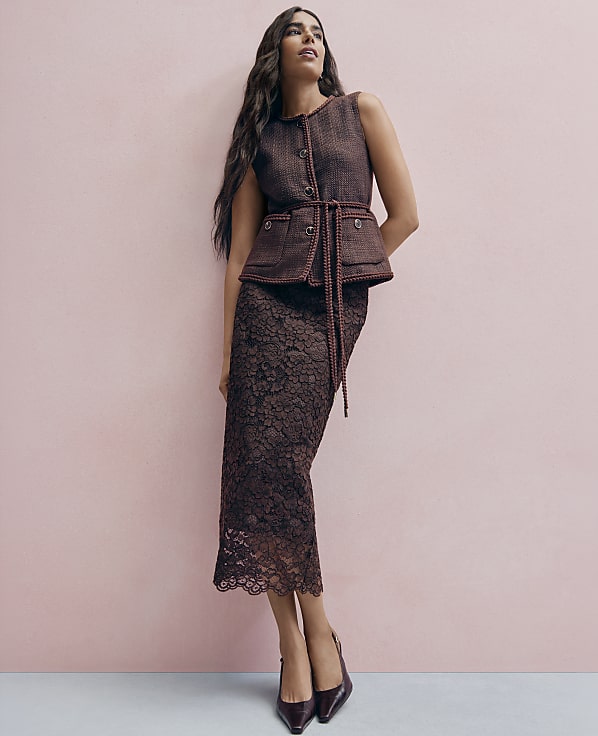 A woman wearing a brown tweed vest and lace skirt stands against a solid pink wall.