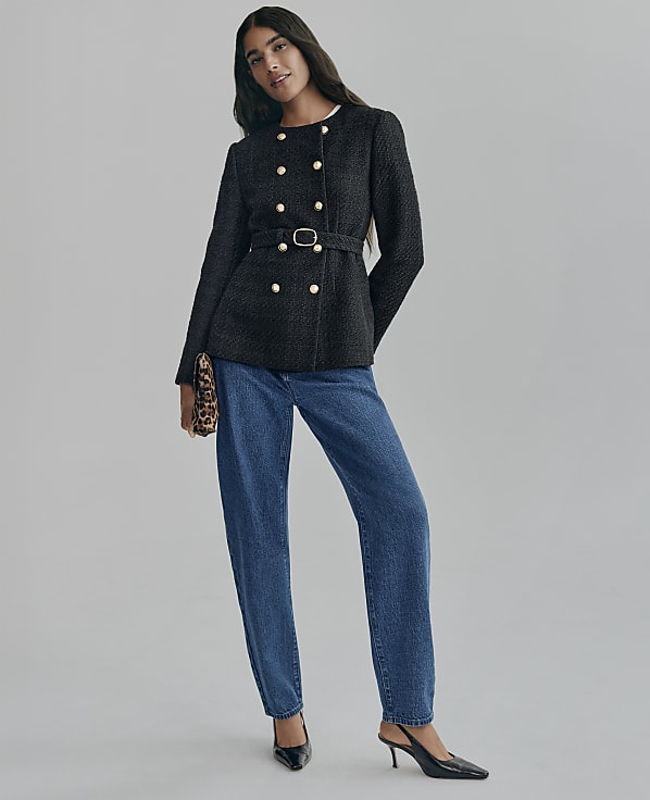 A woman wearing a belted black double-breasted jacket and blue jeans stands against a plain grey background.