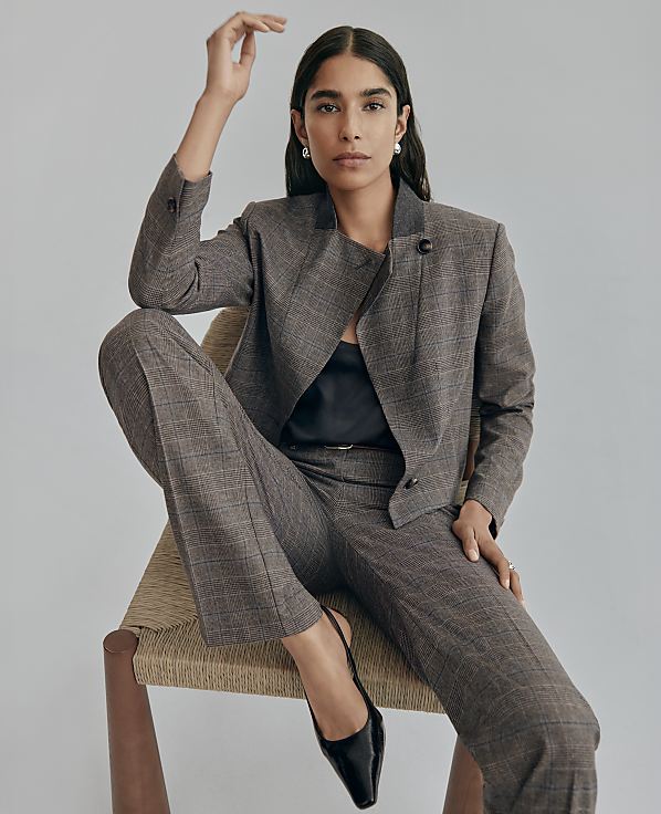 A woman wearing a brown plaid suit and black heels sits in a woven chair against a plain light gray background.