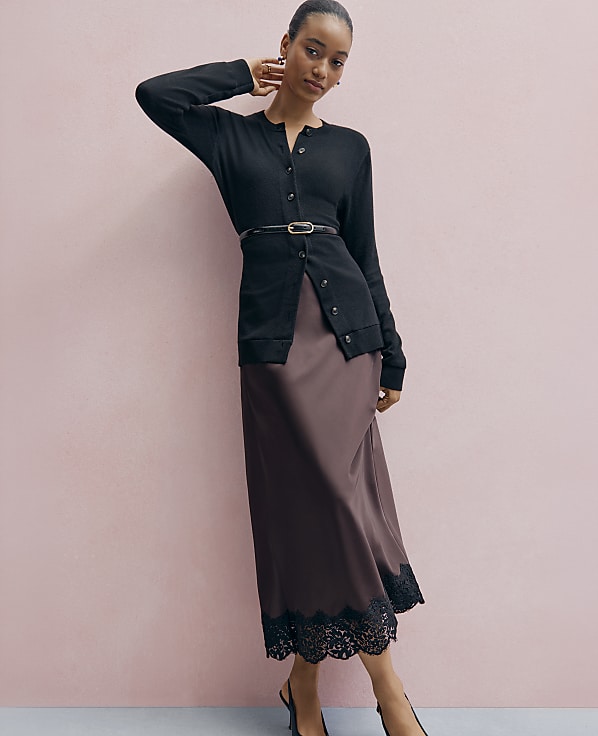A woman wearing a black cardigan and a long brown satin skirt with lace trim stands against a plain pink wall.