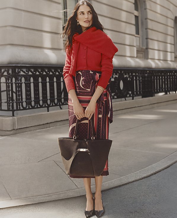 A woman wearing a red cardigan and a patterned skirt stands on a city sidewalk in front of a stone building with a black wrought-iron fence.