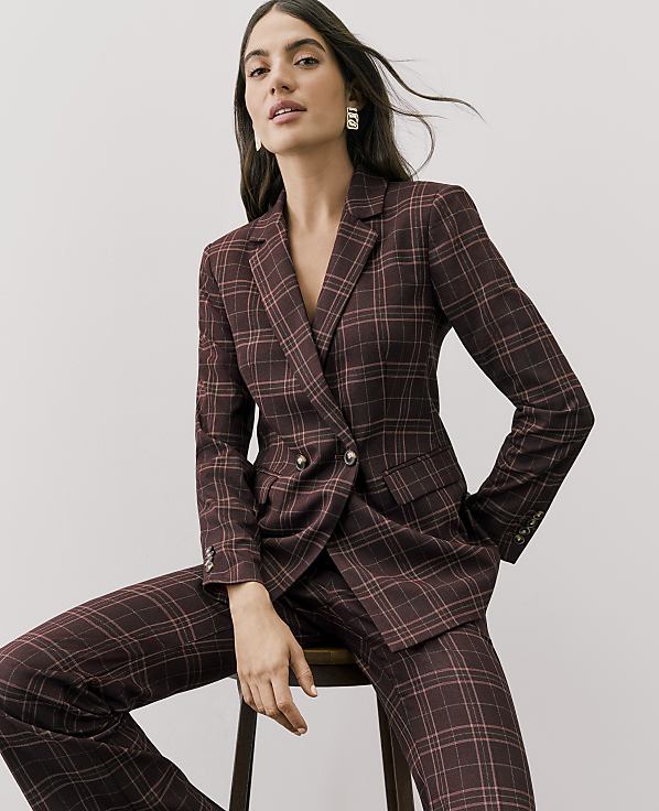 A woman wearing a maroon plaid suit sits on a wooden stool against a plain light gray background.