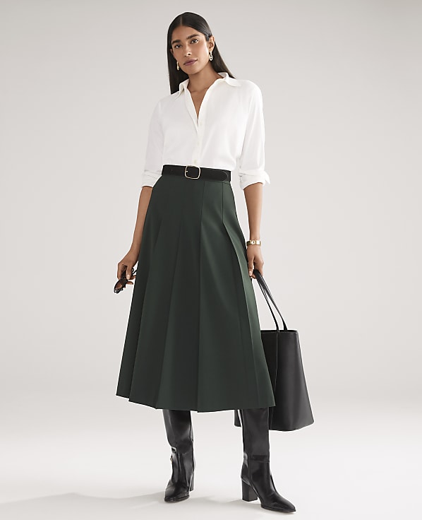A woman wearing a white button-down shirt, a dark green pleated midi skirt, and black boots stands against a plain white background.
