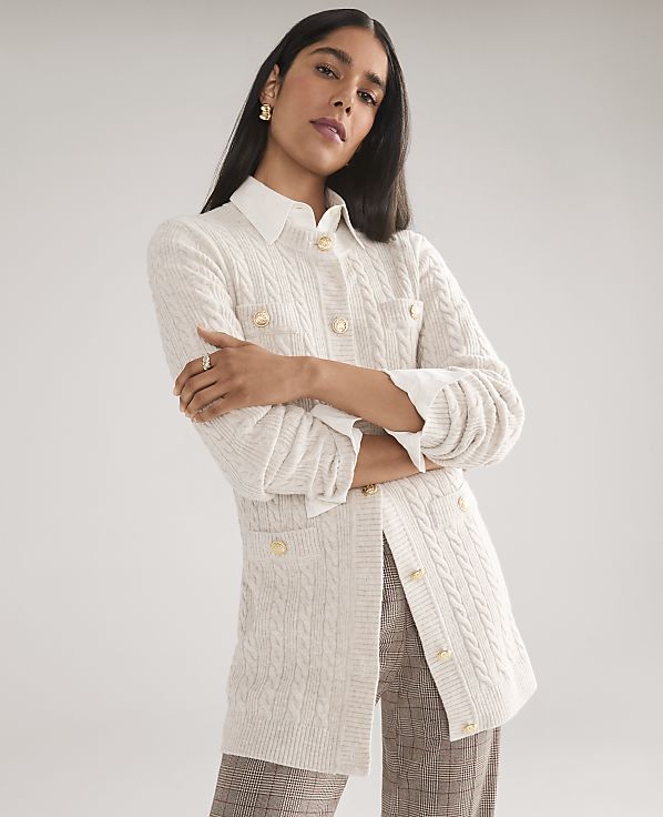 A woman wearing a white cable-knit cardigan over a collared shirt and plaid trousers poses against a plain light gray background.