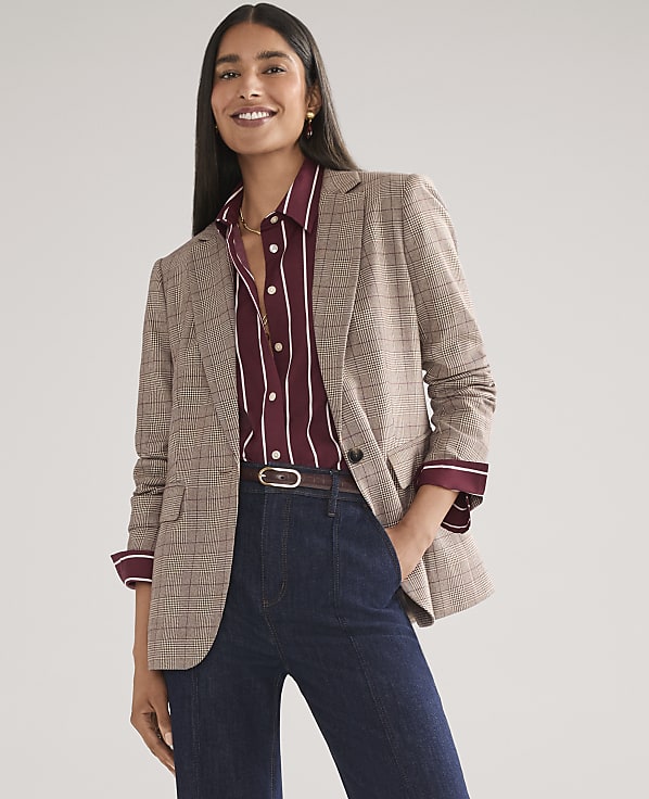 A smiling woman wearing a plaid blazer, striped maroon shirt, and dark blue jeans stands against a plain light gray background.