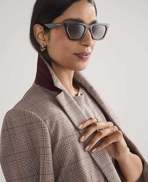 A woman wearing dark sunglasses and a plaid blazer stands against a plain, light-colored background.