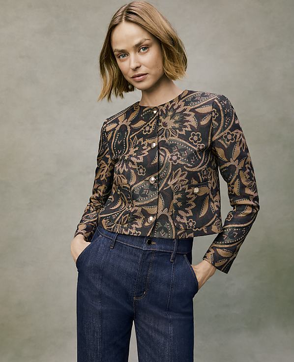 A woman wearing a patterned brown and black jacket and dark blue jeans stands against a neutral, textured grey background.