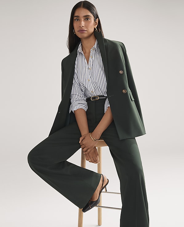 A woman wearing a dark green power suit and a striped shirt stands against a plain white background.
