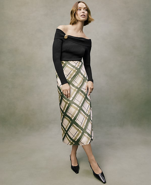 A woman wearing a black off-the-shoulder top and a plaid midi skirt stands against a muted green textured background.