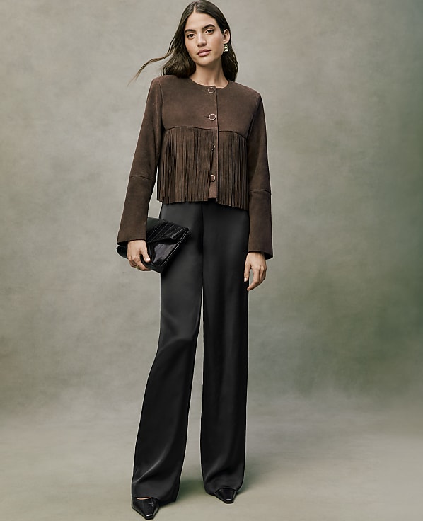 A woman wearing a brown fringed jacket and wide-leg black trousers stands against a neutral, textured grey background.