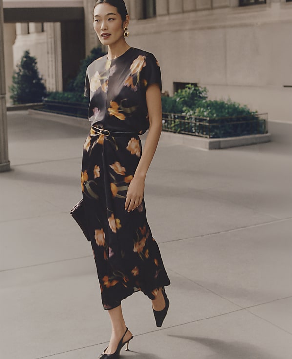 A woman wearing a floral black dress and heels walks along a paved sidewalk in front of a building with greenery.