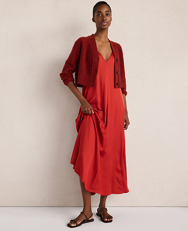 A woman wearing a red satin dress and a matching red cardigan stands against a plain, neutral-toned wall.