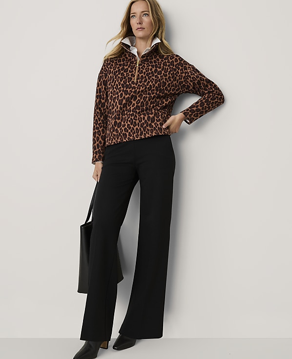 A woman wearing a leopard-print top and black wide-leg trousers stands against a plain white wall.