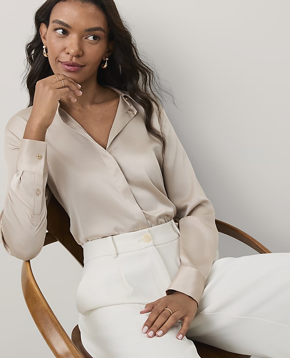 A woman wearing a champagne-colored satin blouse and white trousers sits in a wooden chair against a plain light gray background.