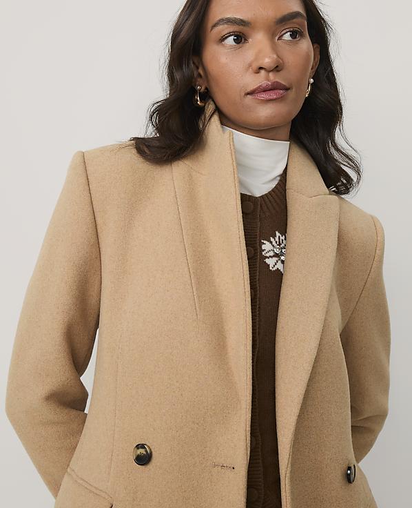 A woman wearing a tan coat over a brown sweater and white turtleneck stands against a plain, light-colored background.