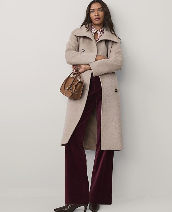 A woman wearing a long beige coat and burgundy velvet trousers stands against a plain white background.