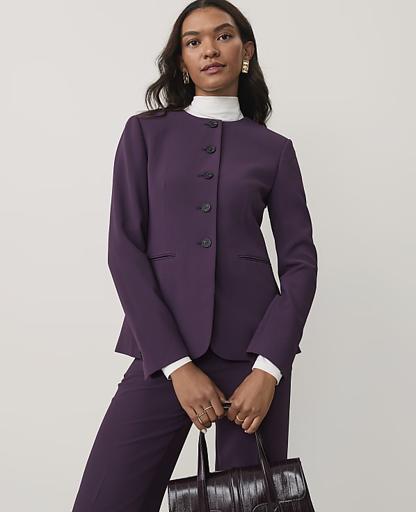 A woman wearing a purple suit and white turtleneck holds a dark burgundy handbag against a plain light gray background.
