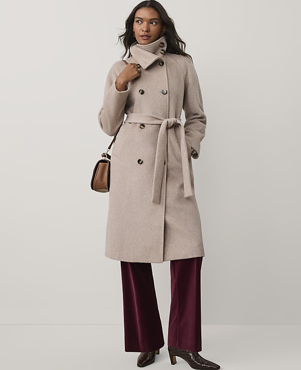 A woman wearing a beige belted coat and burgundy velvet trousers stands against a plain white background.