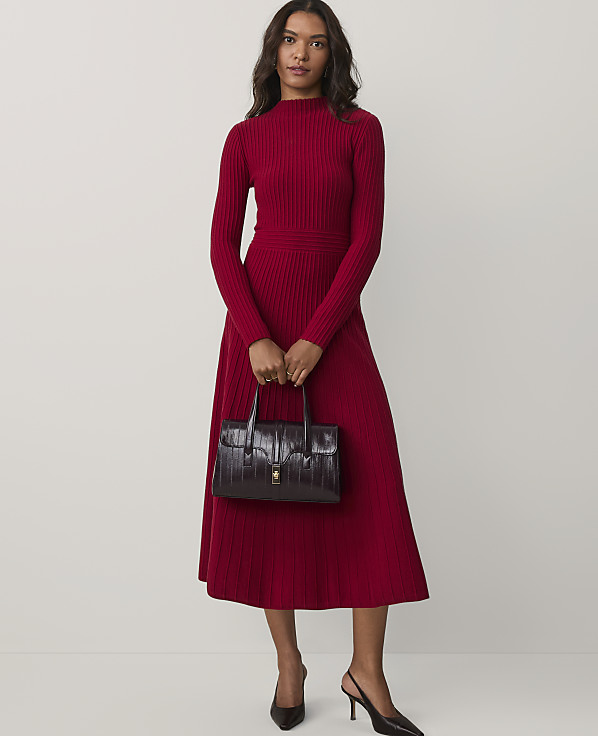 A woman wearing a long-sleeved red ribbed dress and black heels holds a dark burgundy handbag against a plain white background.