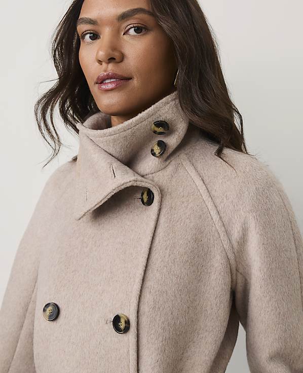 A woman with long brown hair wears a beige wool coat against a plain, light-colored background.