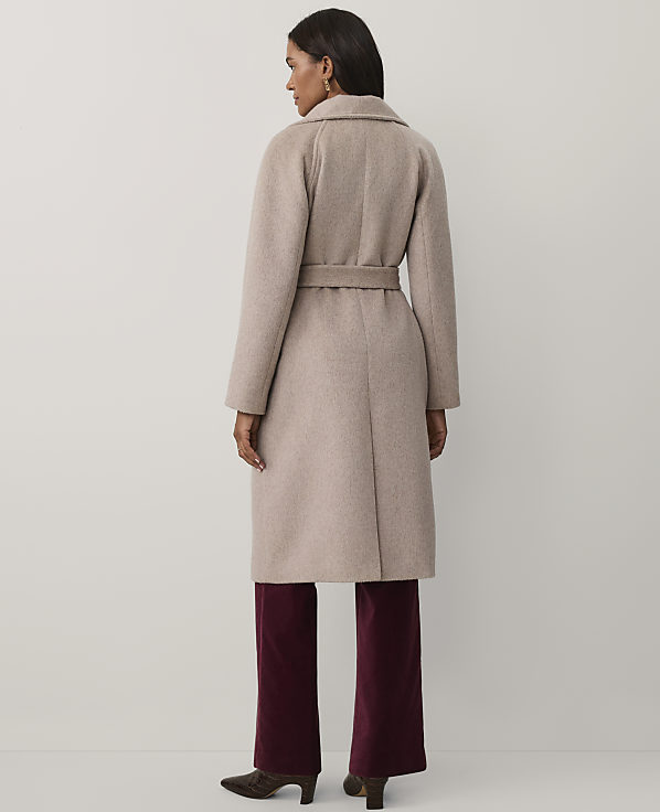A woman wearing a long beige belted coat and burgundy trousers stands with her back to the camera against a plain light grey background.