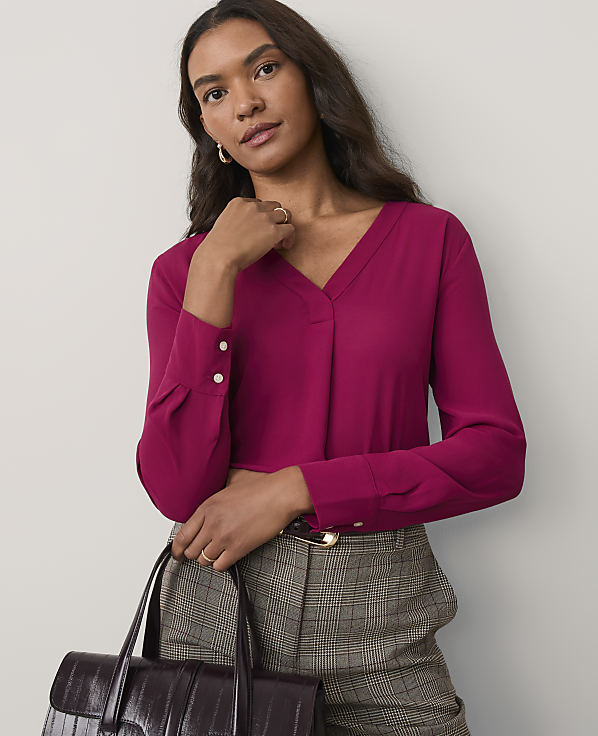 A woman wearing a magenta v-neck blouse and plaid trousers stands against a plain, light-colored wall.