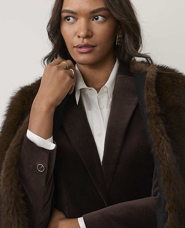 A woman wearing a brown velvet blazer, white shirt, and fur coat poses against a plain, light-colored background.