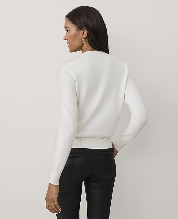 A woman wearing a white sweater and black leather pants stands against a plain, light-colored background.