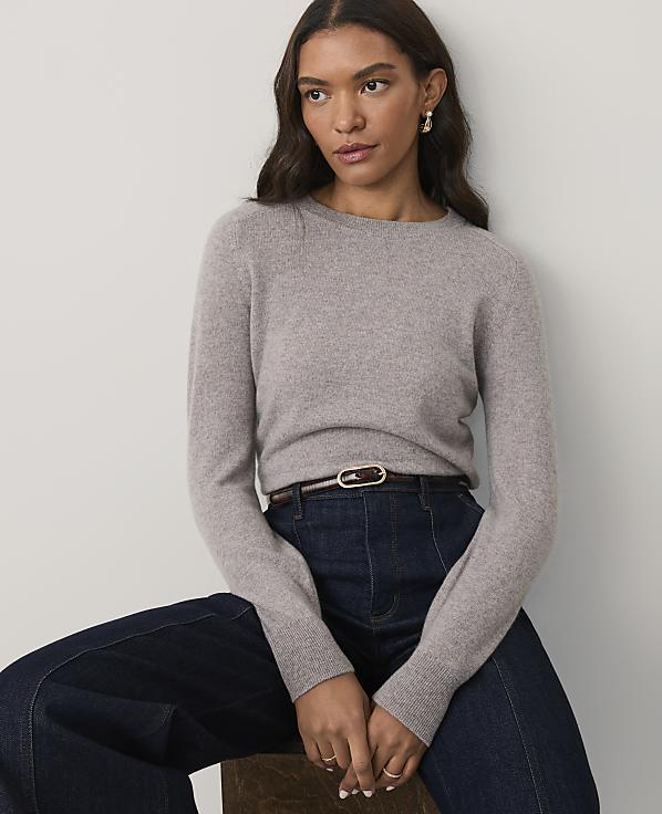 A woman wearing a grey sweater and dark blue jeans sits against a plain white background.