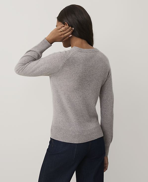 A woman wearing a grey sweater and dark blue jeans stands with her back to the camera against a plain white background.