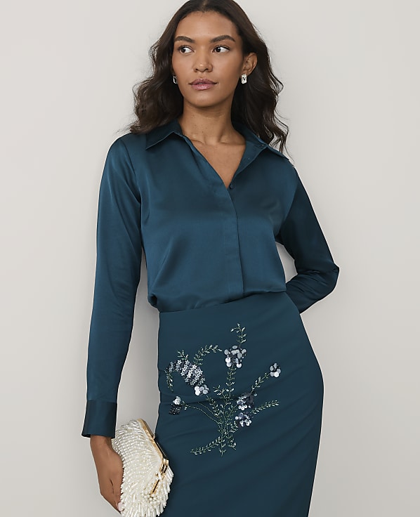 A woman wearing a teal satin blouse and a matching sequined skirt stands against a plain white background.