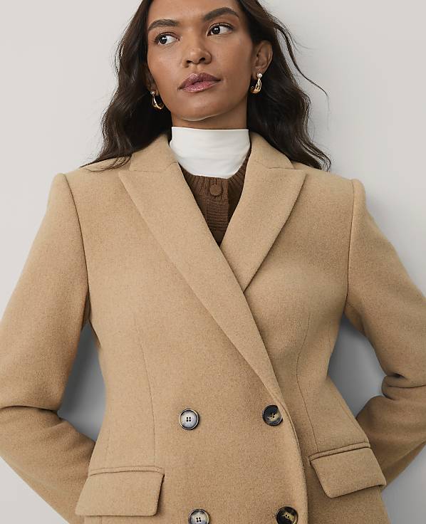 A woman wearing a tan double-breasted blazer over a brown and white layered top poses against a plain light gray background.