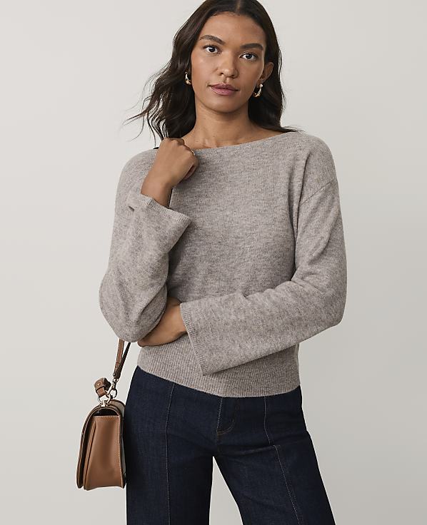 A woman wearing a beige sweater and dark blue jeans stands against a plain light gray background.