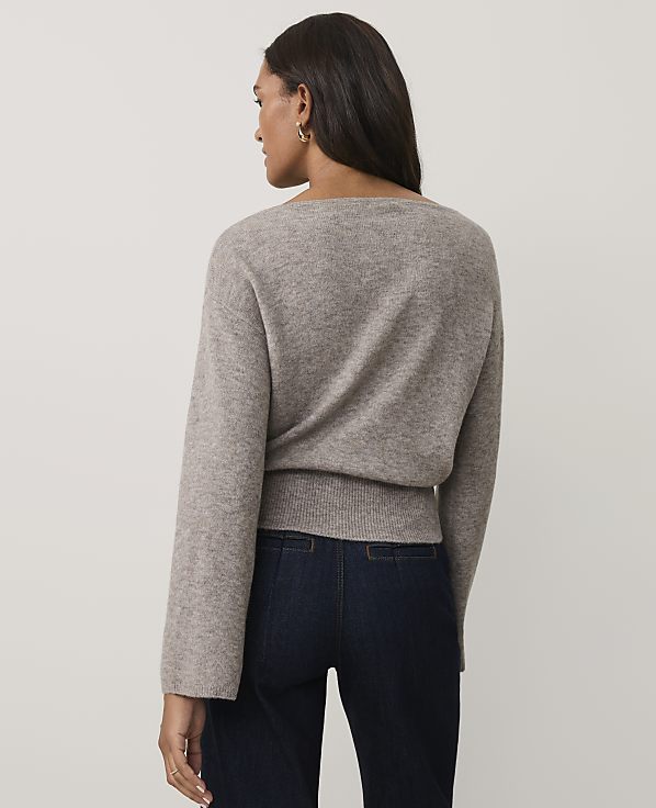 A woman wearing a taupe knit sweater and dark blue jeans stands against a plain light gray background.