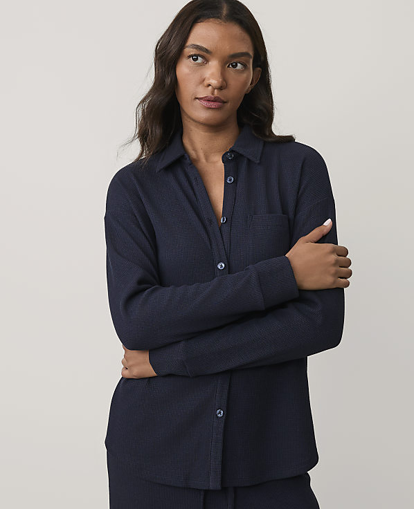 A woman wearing a navy blue ribbed button-down shirt stands with her arms crossed against a plain, light grey background.
