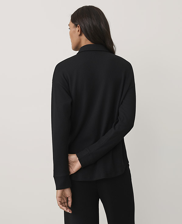 A woman wearing a black long-sleeved ribbed top and black pants stands with her back to the camera against a plain light gray background.