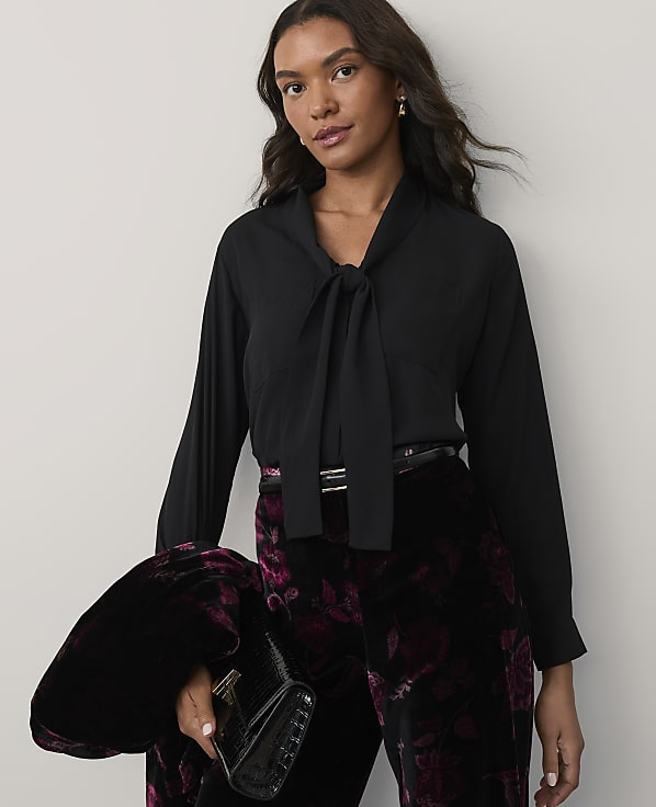 A woman wearing a black tie-neck blouse and floral velvet trousers stands against a plain, light-colored background.