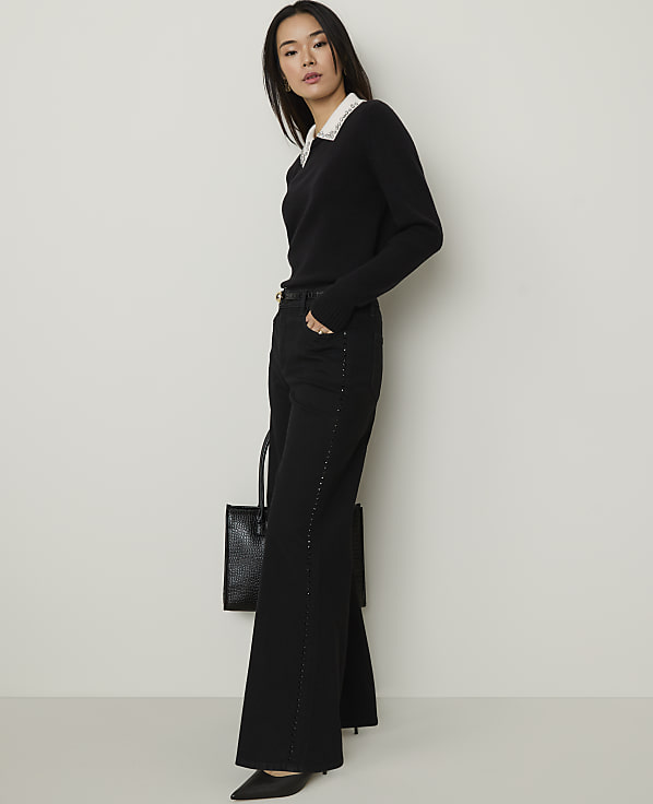 A woman wearing a black sweater with a white collar and wide-leg black trousers stands against a plain, light-colored wall.