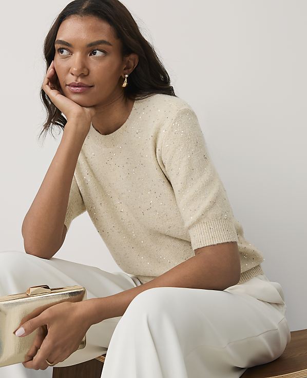 A woman in a shimmering cream sweater and white trousers holds a gold clutch against a plain light gray background.