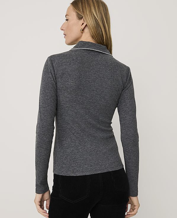 A woman wearing a grey long-sleeved shirt and black pants stands against a plain white background.