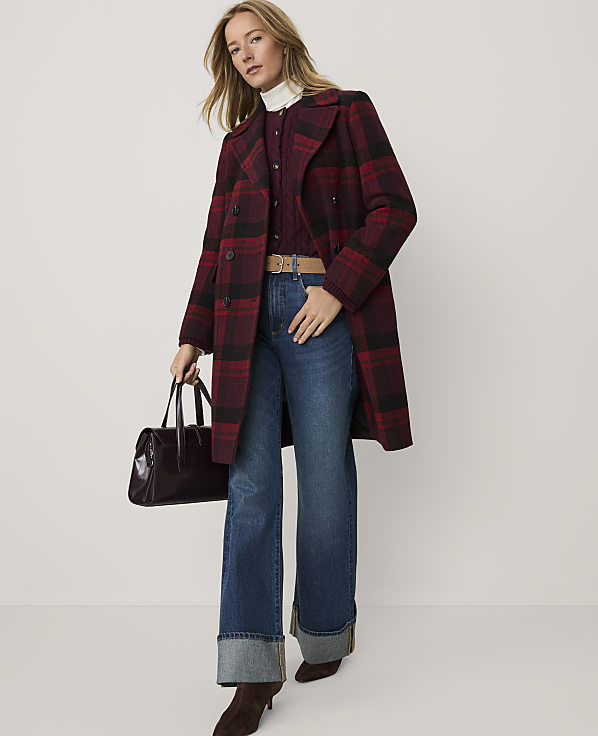 A woman wearing a red plaid coat, blue jeans, and brown boots stands against a plain white background while holding a dark handbag.