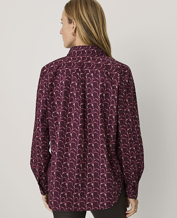 A woman with long blonde hair is seen from behind wearing a maroon patterned button-down shirt against a plain light gray background.