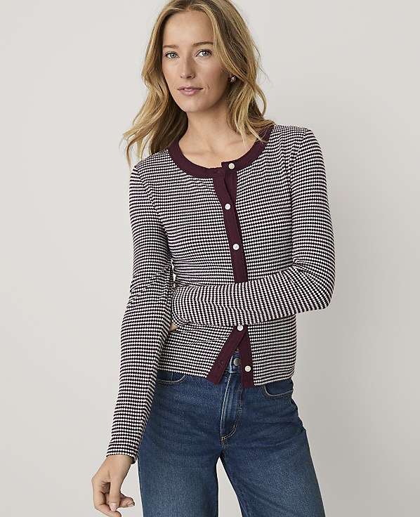 A blonde woman wearing a maroon and white checkered cardigan and blue jeans stands against a plain light gray background.