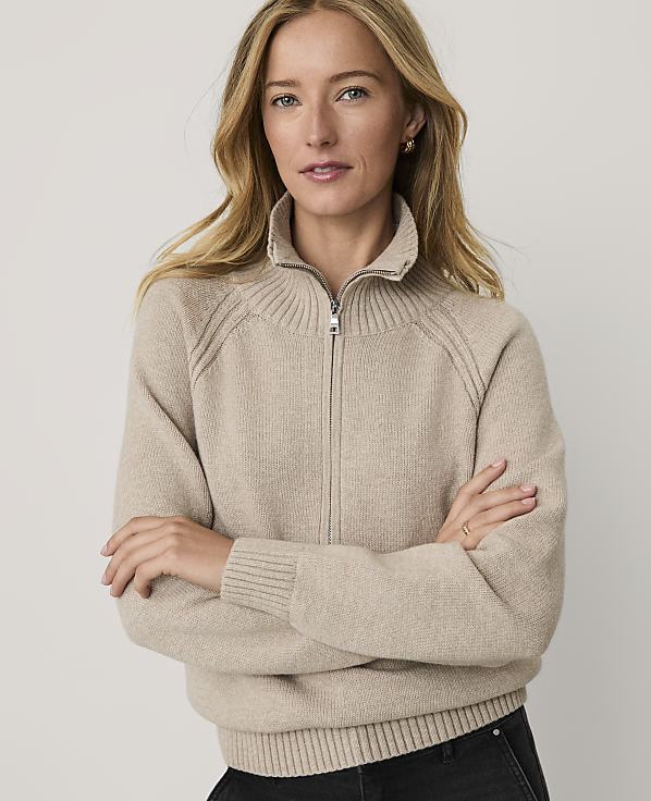 A woman with blonde hair wearing a beige zip-up sweater stands with her arms crossed against a plain, light-colored background.