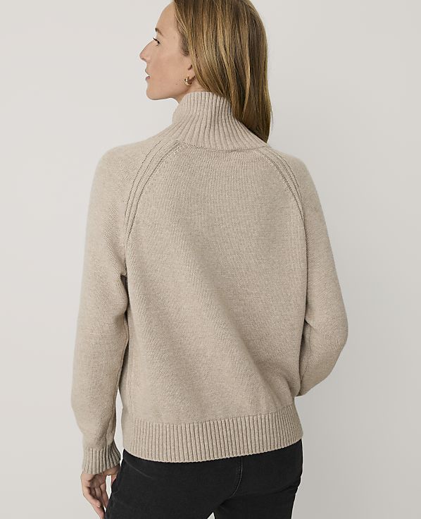 A woman wearing a beige turtleneck sweater and black pants stands against a plain white background.