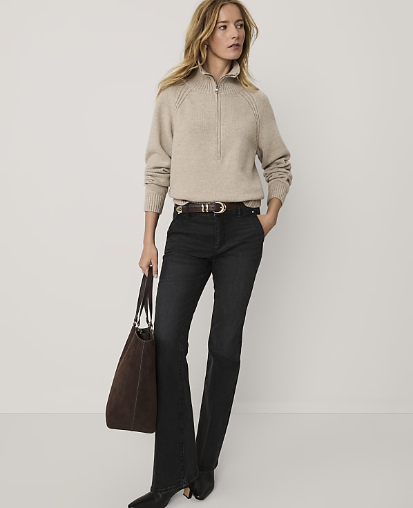 A woman wearing a beige quarter-zip sweater and dark flared jeans stands holding a brown tote bag against a plain white background.